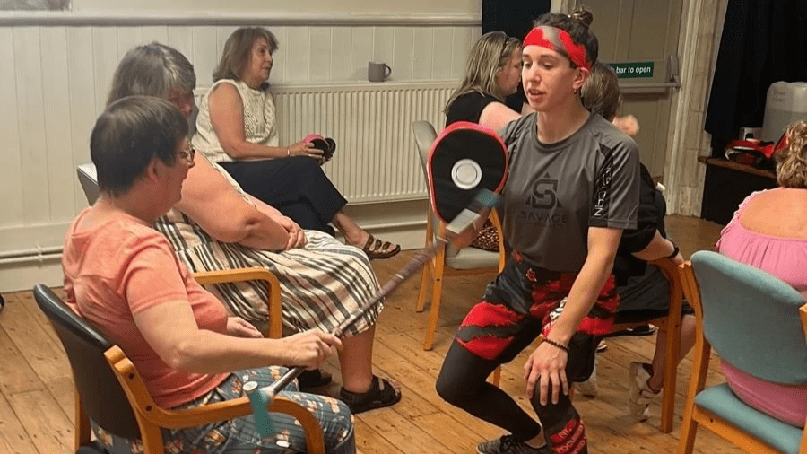 Nikki from Savage Martials Arts is holding a boxing pad, when one of the participants practices hitting it with a walking stick. Everyone is seated around her eagerly awaiting their turn too. The backdrop is Biggin Hall in Dover, where the event was held.