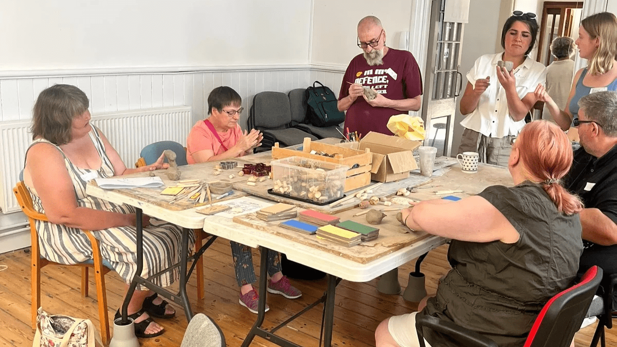 For participants are seated around a table, and three are standing up to make some clay creations. Three people are looking at the pot Rainna has made, she is standing up. The backdrop is Biggin Hall in Dover, where the event was held.