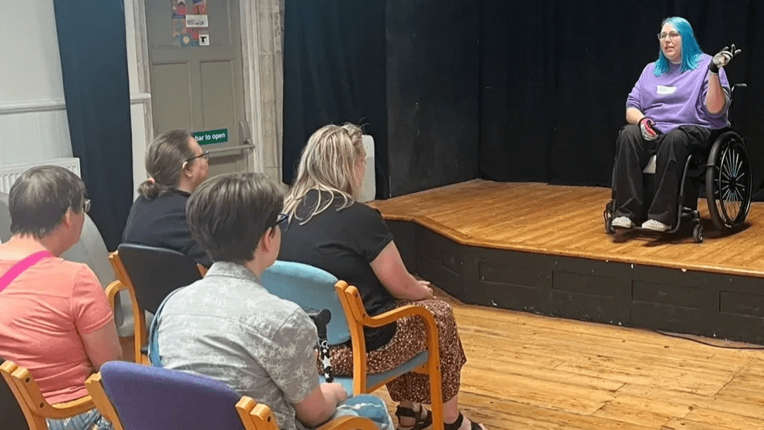 Everyone is sat on chairs with arms looking at me on the stage. I am welcoming everyone and am sat in my wheelchair. The backdrop is Biggin Hall in Dover, where we held our Give it a Go event.