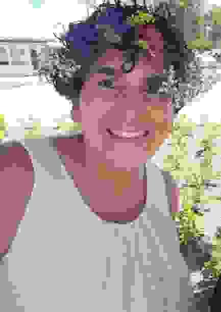 The author.  A smiling very tan woman with short curly hair.  She is wearing a crown of flowers - Baby's breath on either side with purple, yellow and fuschia flowers in the center.  She is wearing a white tank shirt with embroided white flowers.  She is surrounded by shurbs and greenry in the background.