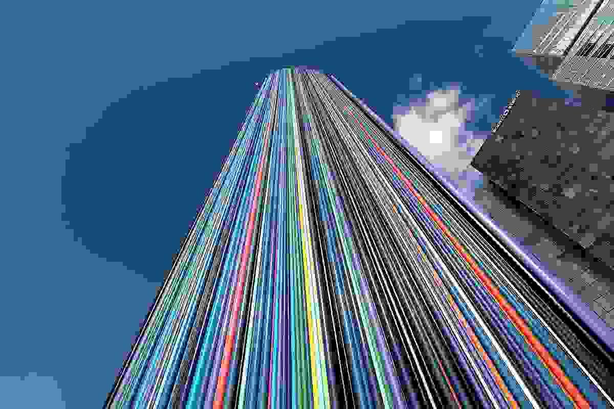 Le Moretti chimney. A chimeny covered in colorful tubes next to some buidlings.  The view is looking up and it's against a blue sky with one cloud