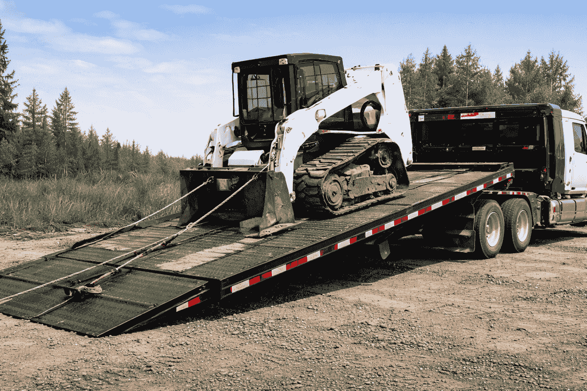 Skid steer being winched onto a tilt-deck trailer during equipment transport. Skid steer being winched onto a tilt-deck trailer during equipment transport.