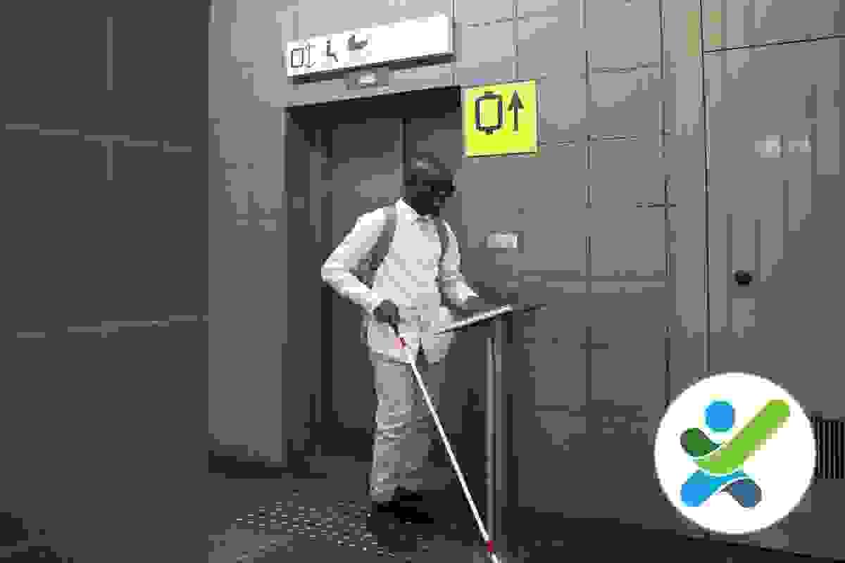 A man with vision impairment is using a long white cane and is standing in front of a lift doorway and is operating a destination control system panel A man with vision impairment is using a long white cane and is standing in front of a lift doorway and is operating a destination control system panel