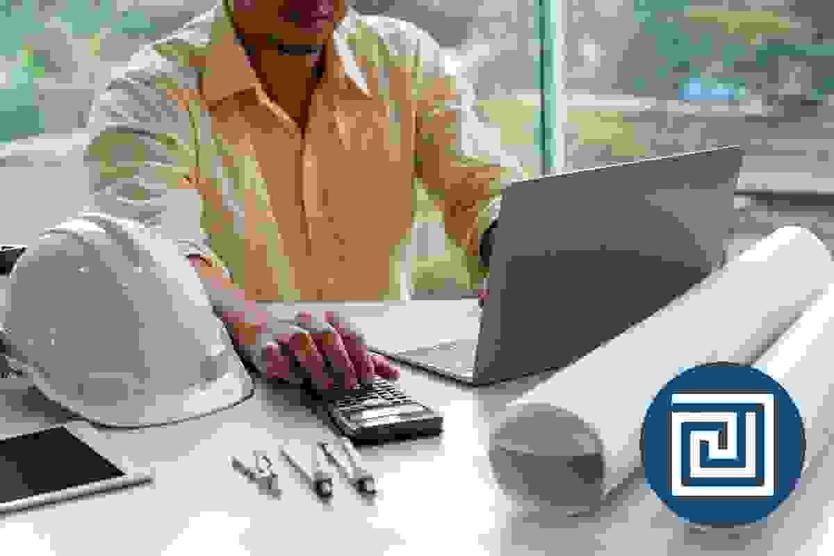 A male architect is looking at a laptop, while using a calculator to check a ramp gradient, there are architectural drawings and a hardhat on the desk A male architect is looking at a laptop, while using a calculator to check a ramp gradient, there are architectural drawings and a hardhat on the desk