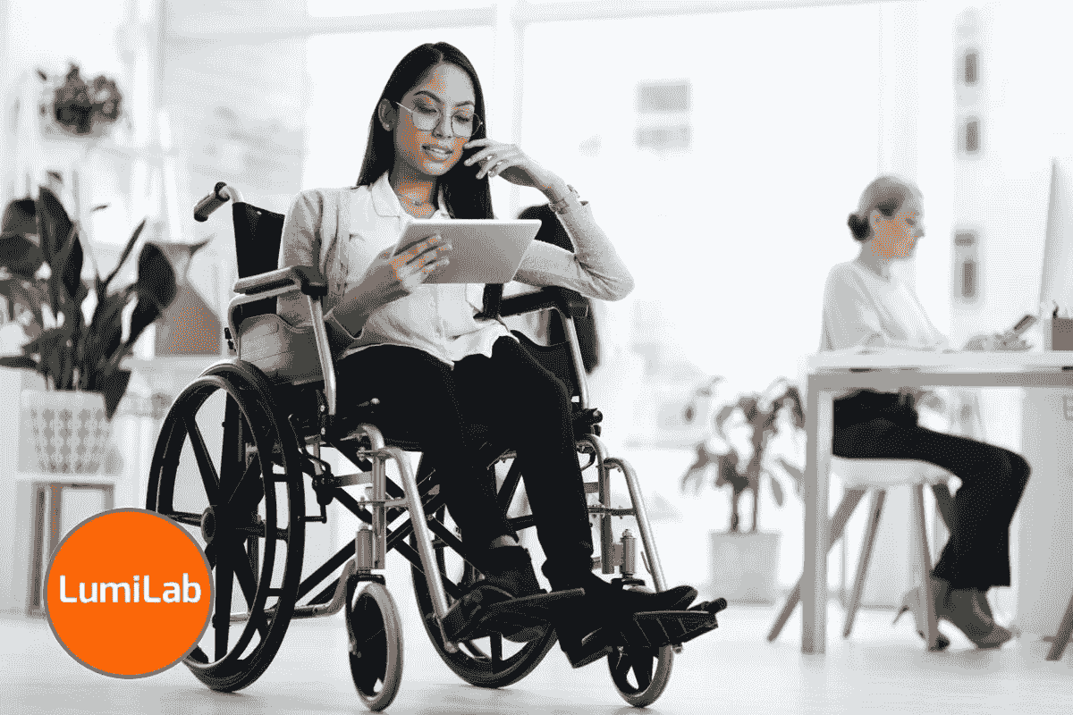 A woman in a wheelchair uses a tablet in an office, with another person working at a desk in the background. A woman in a wheelchair uses a tablet in an office, with another person working at a desk in the background.