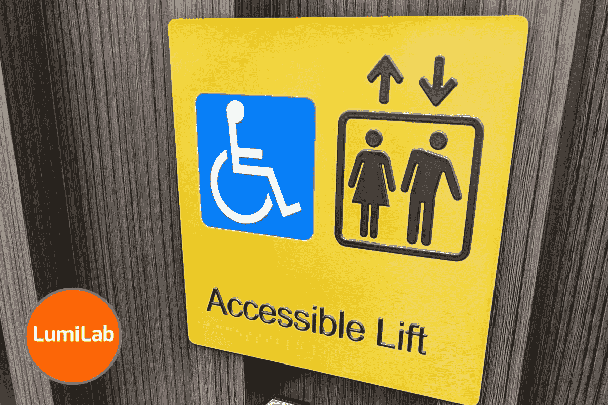 A yellow "Accessible Lift" Braille sign features a wheelchair symbol, elevator icons with a man and woman, and up and down arrows. A yellow "Accessible Lift" Braille sign features a wheelchair symbol, elevator icons with a man and woman, and up and down arrows.