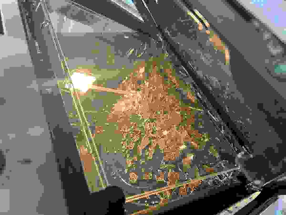 Interior of a oven with shattered glass scattered across the bottom. The broken glass has exploded into small shards. The oven light is on, illuminating the damage. A control dial is partially visible on the left side of the frame.