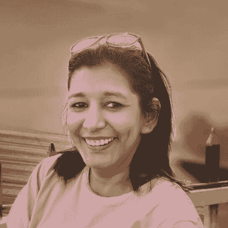 "Portrait of a woman with medium-length dark hair, wearing a pink top, smiling brightly while looking sideways at the camera. She is seated on a wooden bench with a warm sunset in the background.