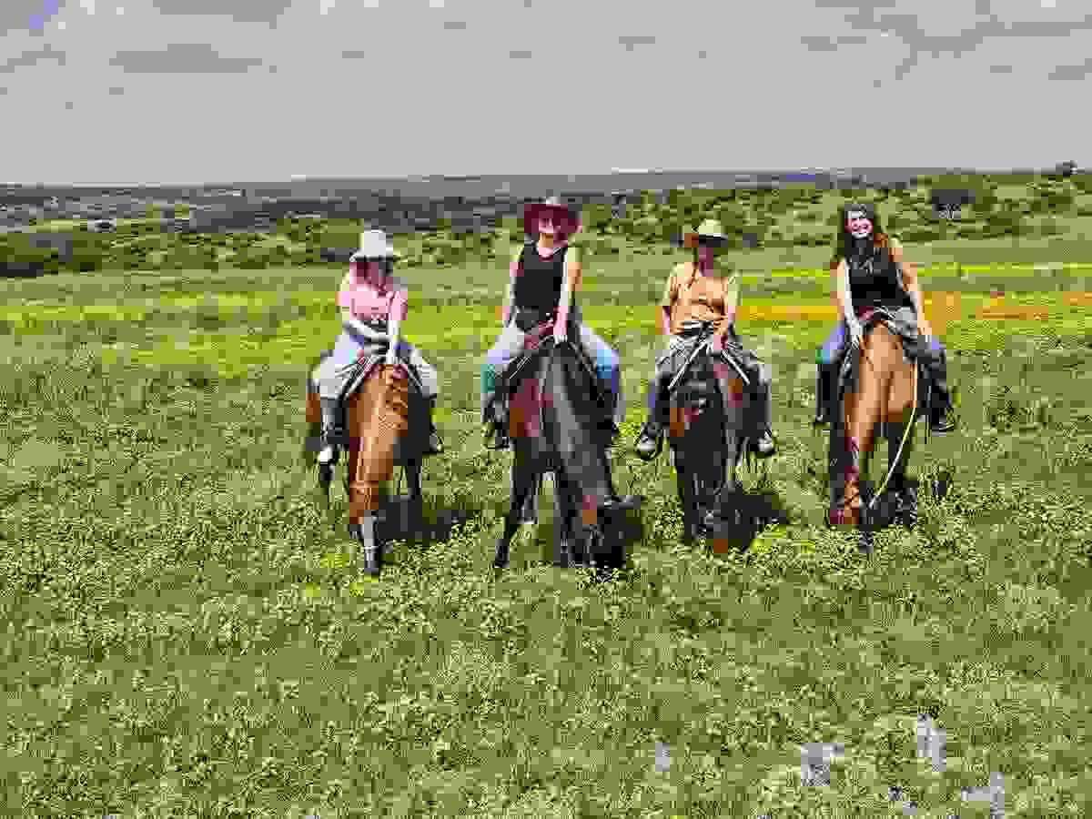 Four women on horseback at Presa De Los Torres near San Miguel de Allende, surrounded by yellow wildflowers, as the horses graze peacefully in the lush green landscape during rainy season.