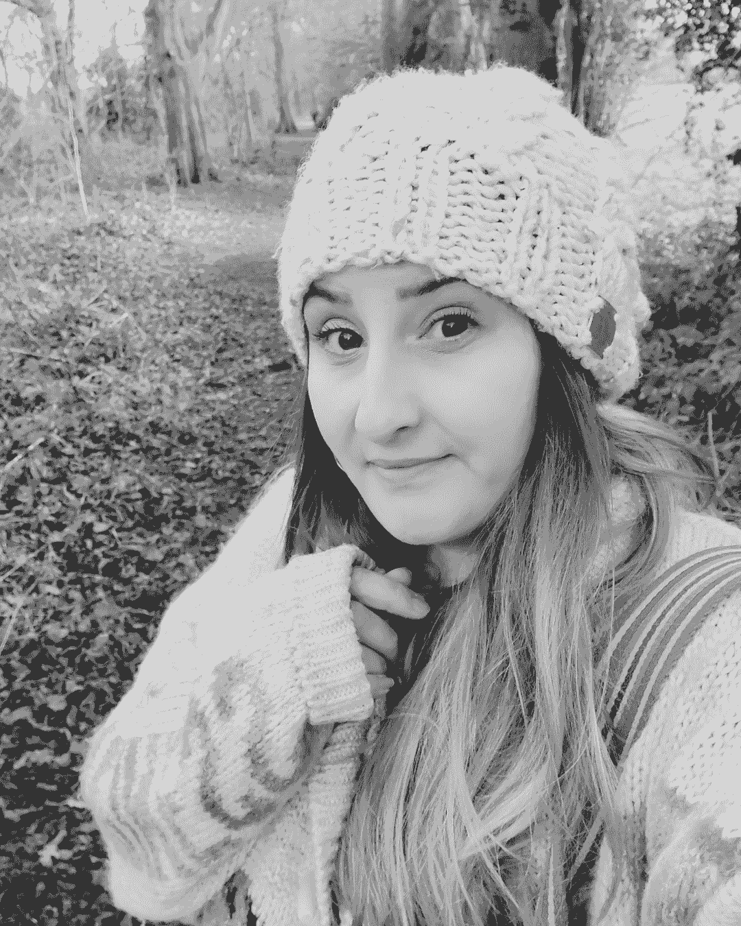 A black and white photograph of Charlie Edwards, a white woman in her thirties. Charlie stands in the woodland, with a leaf-covered path and trees behind her. She wears a light coloured jumper, pulled down over one hand, and a white woollen hat. Her brown hair falls around her shoulders. A black and white photograph of Charlie Edwards, a white woman in her thirties. Charlie stands in the woodland, with a leaf-covered path and trees behind her. She wears a light coloured jumper, pulled down over one hand, and a white woollen hat. Her brown hair falls around her shoulders.