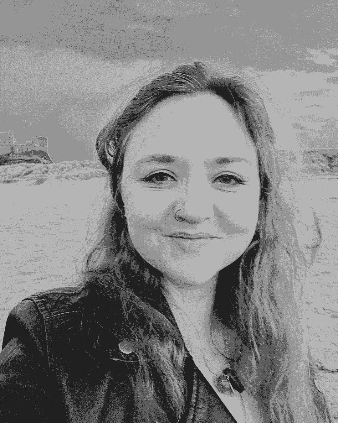 A black and white photograph of Ceryn Rowntree, a white woman in her 40s. Ceryn stands on a beach with the sky and a castle (Bamburgh in Northumberland) behind her. She wears a black jacket and has long, wavy dark hair over her shoulders. A black and white photograph of Ceryn Rowntree, a white woman in her 40s. Ceryn stands on a beach with the sky and a castle (Bamburgh in Northumberland) behind her. She wears a black jacket and has long, wavy dark hair over her shoulders.