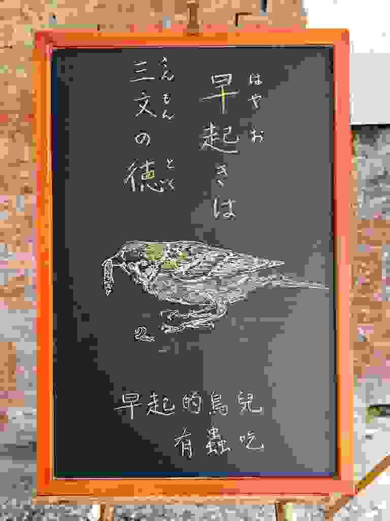 小さい黒板にチョークで絵が描いてあります。日本語のことわざ・慣用表現を紹介するための絵です。上部に縦書き二行で「早起きは／三文の徳」と書いてあります。中央少し下に、大きく雀のイラストが描いてあります。雀はくちばしに虫を加えています。下部に、このことわざに相当する中文のことわざ「早起的鳥兒有蟲吃」と書いてあります。これは、早起きの鳥は虫にありつけるという意味です。