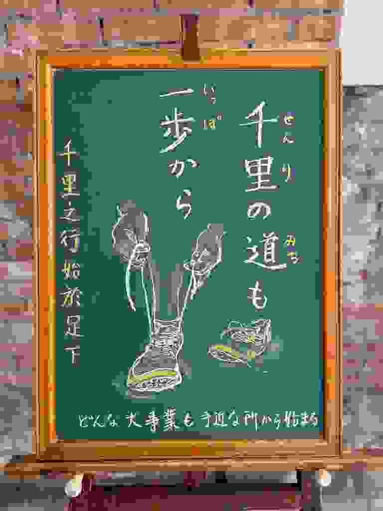 小さい黒板にチョークで絵が描いてあります。日本語のことわざ・慣用表現を紹介するための絵です。縦長の黒板で右上から縦書き二行で「千里の道も/一歩から」と書いてあります。左部分から下部にかけて、ランニングシューズを履いている人の足とシューズの部分のイラストが描いてあります。右足をシューズに入れていて、くつひもを両手でぎゅっと引っ張った瞬間です。左足はまだはいていなくて、そばに左足用のシューズが見えます。