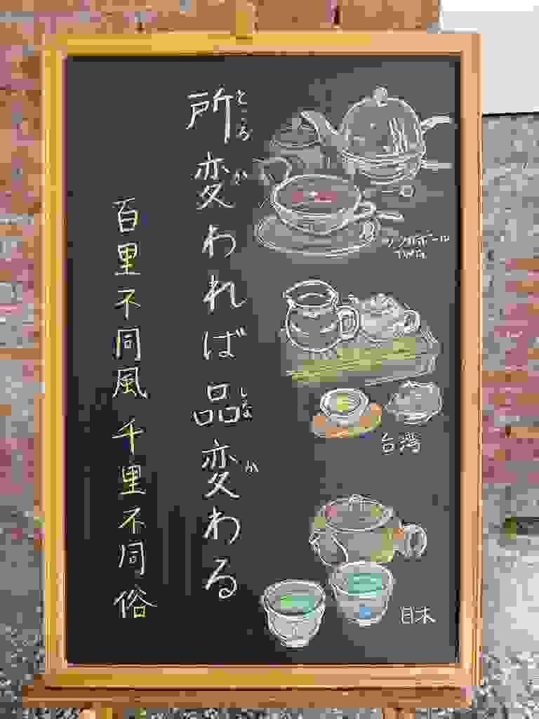 小さい黒板にチョークで絵が描いてあります。日本語のことわざ・慣用表現を紹介するための絵です。縦長の黒板で、左側に一行で「所変われば品変わる」と書いてあり、その左にこのことわざに相当する中国語の表現「百里不同風、千里不同俗」と書いてあります。 右側には、お茶の道具のイラストが三種類描いてあります。 上から紅茶のティーポットと、白いティーカップで、紅茶が注いであります。カップにはTWGという文字が見えます。シンガポールのTWGのカップです。 その下は、台湾のお茶セットにイラストです。木製の茶台の上にガラス製の茶海(ピッチャー)と白い陶器の小ぶりの茶壷(急須)のイラストが描いてあります。手前に茶杯があり、黄緑のお茶が注いであります。 一番下には日本茶の急須と湯呑2つのイラストが描いてあります。湯呑には緑茶が注いであります。