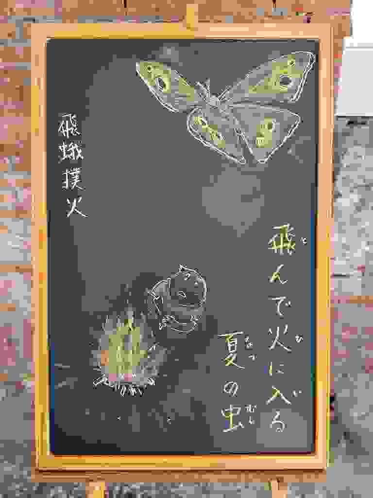 小さい黒板にチョークで絵が描いてあります。日本語のことわざ・慣用表現を紹介するための絵です。右下に縦書き二行で「飛んで火に入る／夏の虫」と書いてあります。その上部には、大きな蛾が羽を広げて飛んでいるイラストが描いてあります。左下には、男性が焚き火をしているイラストが、空からみた視点で描いてあります。大きな炎が上がり、その側で男性は上を飛ぶ蛾を見上げています。