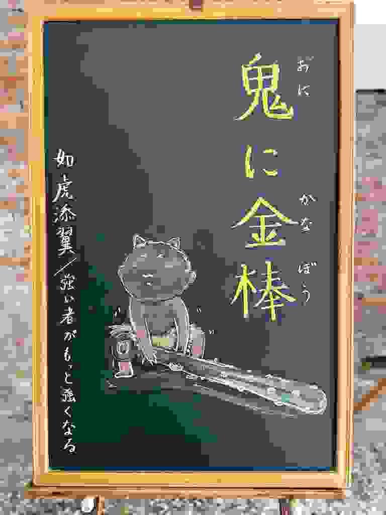 小さい黒板にチョークで絵が描いてあります。日本語のことわざ・慣用表現を紹介するための絵です。縦長の黒板で右側に縦書きで「鬼に金棒」と書いてあります。左下に、赤鬼の子どもが大きな金棒を持ち上げようとして力を入れているイラストが描いてあります。目をつぶり、上を向いて中腰で力をこめていますが、あまり持ち上がっていません