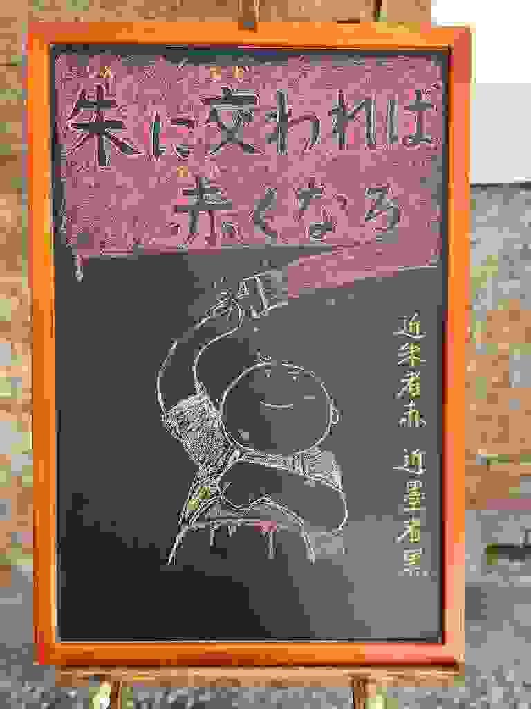 小さい黒板にチョークで絵が描いてあります。日本語のことわざ・慣用表現を紹介するための絵です。上部に横書き２行で「朱に交われば／赤くなる」と書いてあります。背景は赤で文字の部分に色はなく黒板の地の色が見えています。 下部に赤いペンキを塗っている男の子のイラストが描いてあります。右手に刷毛を持って上部を勢いよく塗っていて、もう一方の手でペンキの缶を前に持っています。