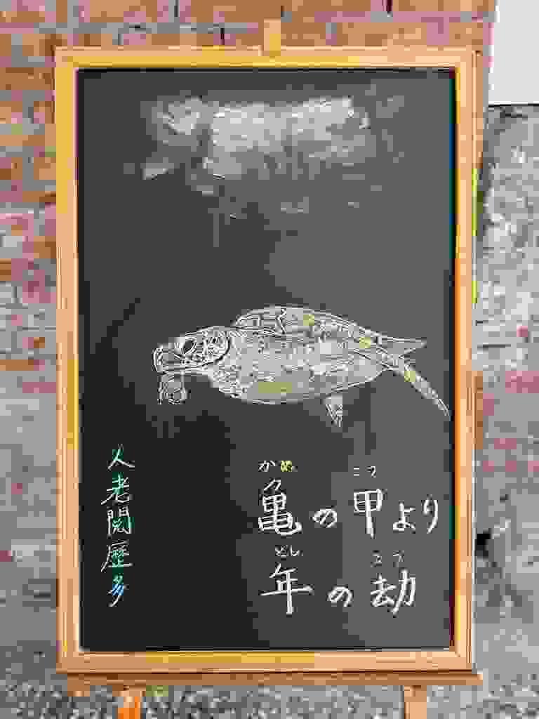 小さい黒板にチョークで絵が描いてあります。日本語のことわざ・慣用表現を紹介するための絵です。縦長の黒板で中央に海の中を泳ぐウミガメのイラストが描いてあります。その上に水面らしき部分が光を浴びて白くなっています。ウミガメの下に横書き二行で「亀の甲より/年の劫」と書いてあります。左下に、このことわざの説明にあたる中国語が縦書きで小さめに書いてあります「人老閱歷多」
