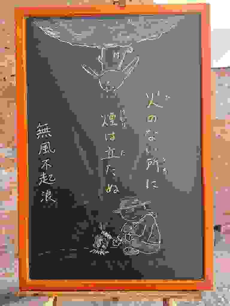 小さい黒板にチョークで絵が描いてあります。日本語のことわざ・慣用表現を紹介するための絵です。縦長の黒板で中央に二行で「火のない所に／煙は立たぬ」と書いてあり、下部にキャンプで焚き火をしている人のイラストが描いてあります。帽子を被り、長袖のシャツで地面に直接座り込んでいます。手に串に刺した肉(のような物)を持っていて焚き火の上にかざしています。そこから、白い煙が上部に上がっています。上部には月とおぼしき球体の一部があり、そこに立ったウサギが下から上がってきた煙に気づいた様子のイラストが描いてあります。