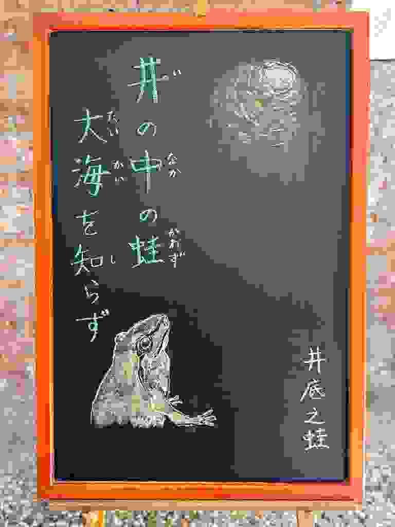 小さい黒板にチョークで絵が描いてあります。日本語のことわざ・慣用表現を紹介するための絵です。縦長の黒板で左上に縦書き二行で「井の中の蛙／大海を知らず」と書いてあります。その下に緑のカエルが上を見上げているイラストが描いてあります。カエルが見上げるのその先に井戸の入口のイラストがあります。レンガで丸く組まれている構造が外からの光で見えています。