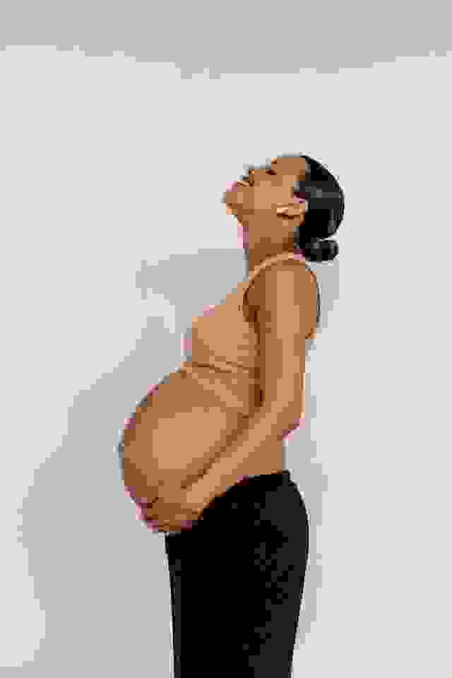 A third-trimester woman of colour holds her bump with both hands, smiling and looking upwards. A third-trimester woman of colour holds her bump with both hands, smiling and looking upwards.
