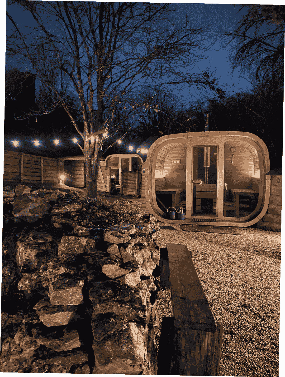 Two saunas glow against a dark winter sky. A bare tree's braches are lit with a string of lightbulbs. Two saunas glow against a dark winter sky. A bare tree's braches are lit with a string of lightbulbs.
