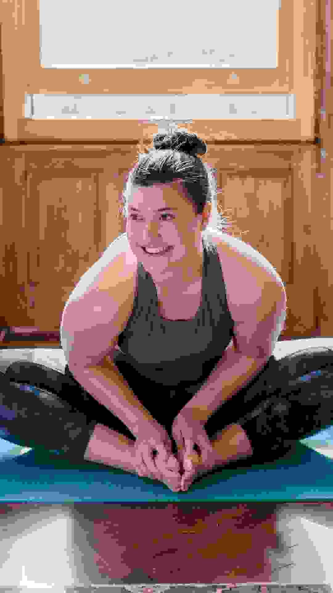 Female client sits smiling on a blue yoga mat. Her feet are togeher, knees bent out to the side in a butterfly yoga pose. Female client sits smiling on a blue yoga mat. Her feet are togeher, knees bent out to the side in a butterfly yoga pose.