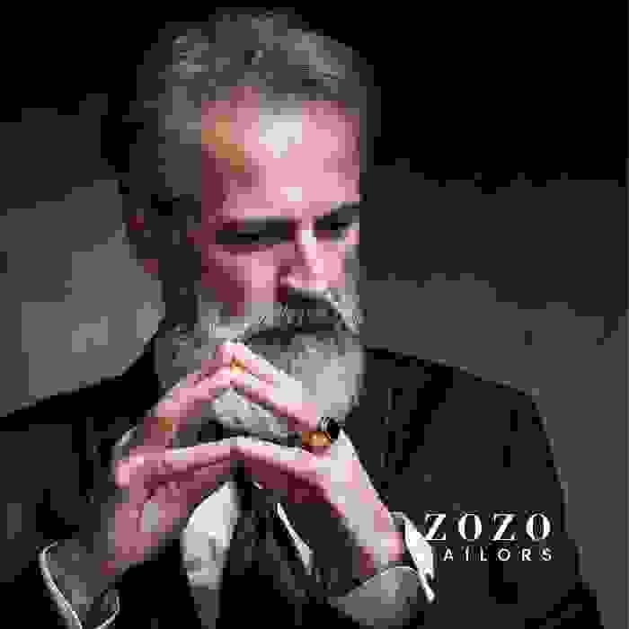 image of sartorial man, in close up, showing dark suit with white shirt and dark blue tie, he is holding his hands together in a contemplative pose