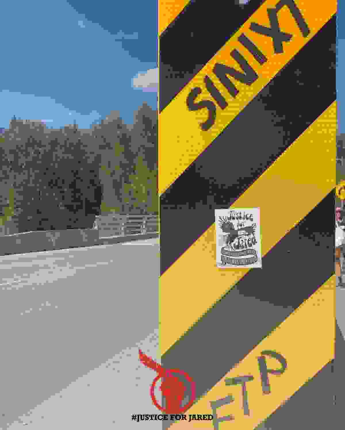 A graffitied roadside sign (black and yellow diagonal stripes) with the text “SINIXT” and “FTP.” A black and white Justice For Jared sticker is stuck in the centre. At the bottom of the graphic is the Justice For Jared logo, a red fist holding up an eagle feather.