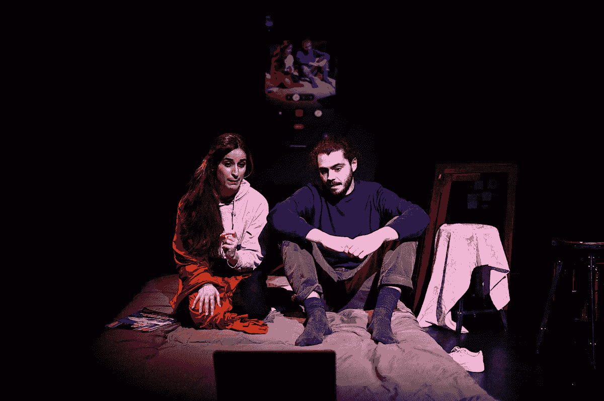 Un couple est assis côte à côte sur un lit devant un écran d'ordinateur. Ce sont des personnages de la pièce Lueurs. Un couple est assis côte à côte sur un lit devant un écran d'ordinateur. Ce sont des personnages de la pièce Lueurs.