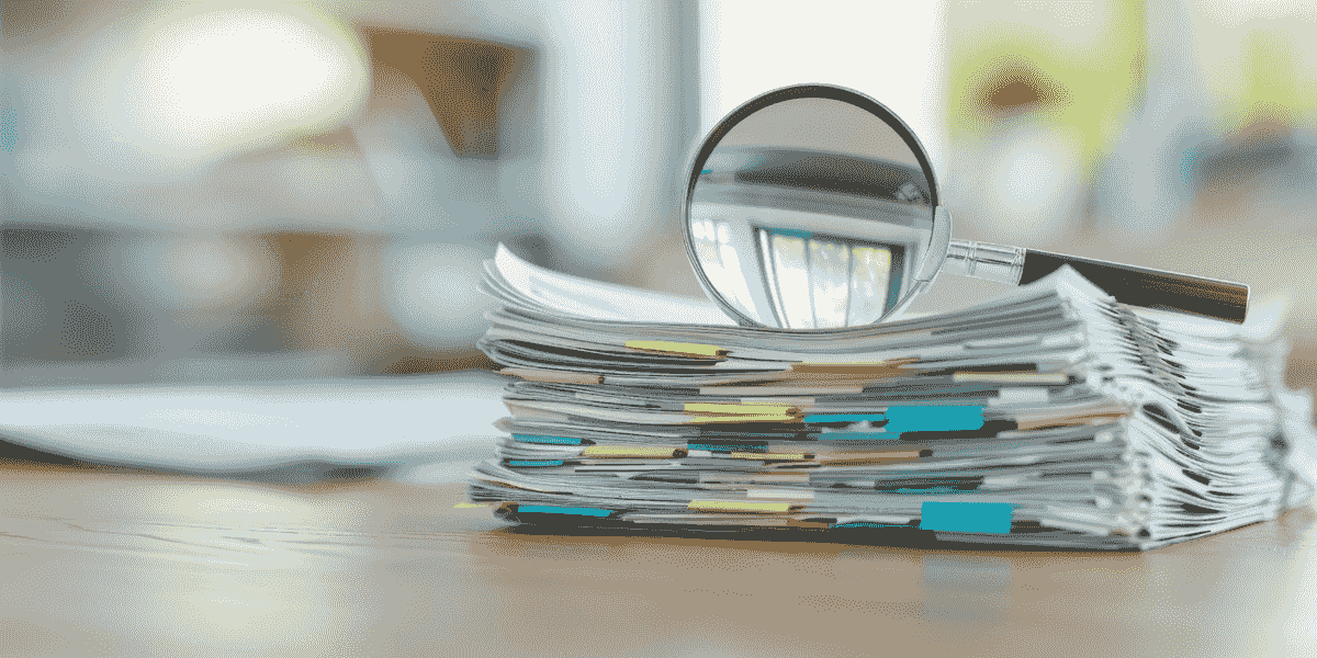 Une loupe est posée sur une grande pile de documents avec des trombones de couleur, sur un bureau en bois.