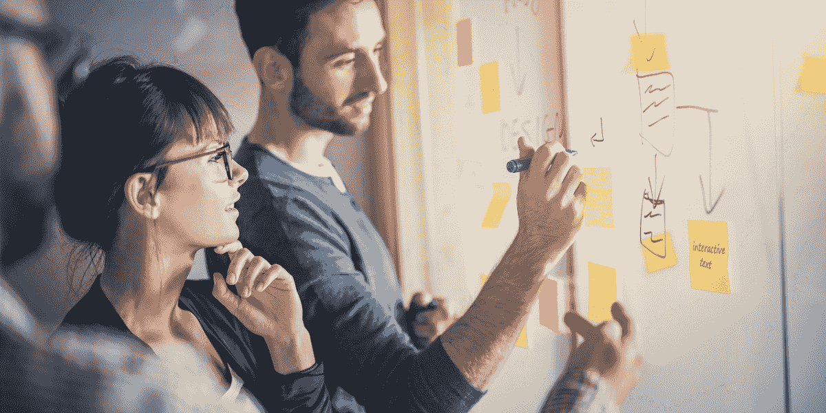 Trois personnes collaborent devant un tableau blanc couvert de notes autocollantes et de diagrammes, une personne écrit tandis que les autres observent et discutent d’idées.