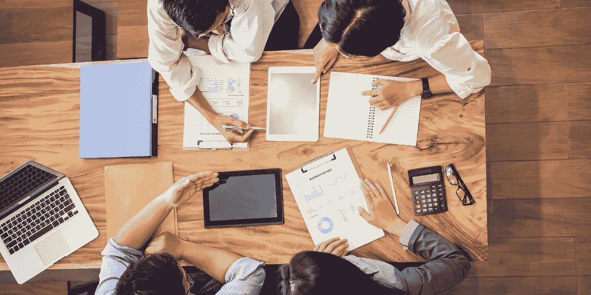 Vue aérienne de professionnels divers collaborant autour d’une table en bois, avec des ordinateurs portables, des rapports, des tablettes et des documents.