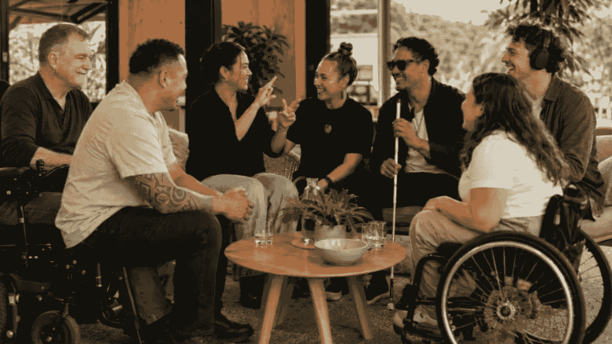 Pip, seated in a power wheelchair, joins six diverse people in a lively conversation around a table. The group includes a person signing, a man holding a white cane, a headphone wearer and a manual wheelchair user. Pip, seated in a power wheelchair, joins six diverse people in a lively conversation around a table. The group includes a person signing, a man holding a white cane, a headphone wearer and a manual wheelchair user.