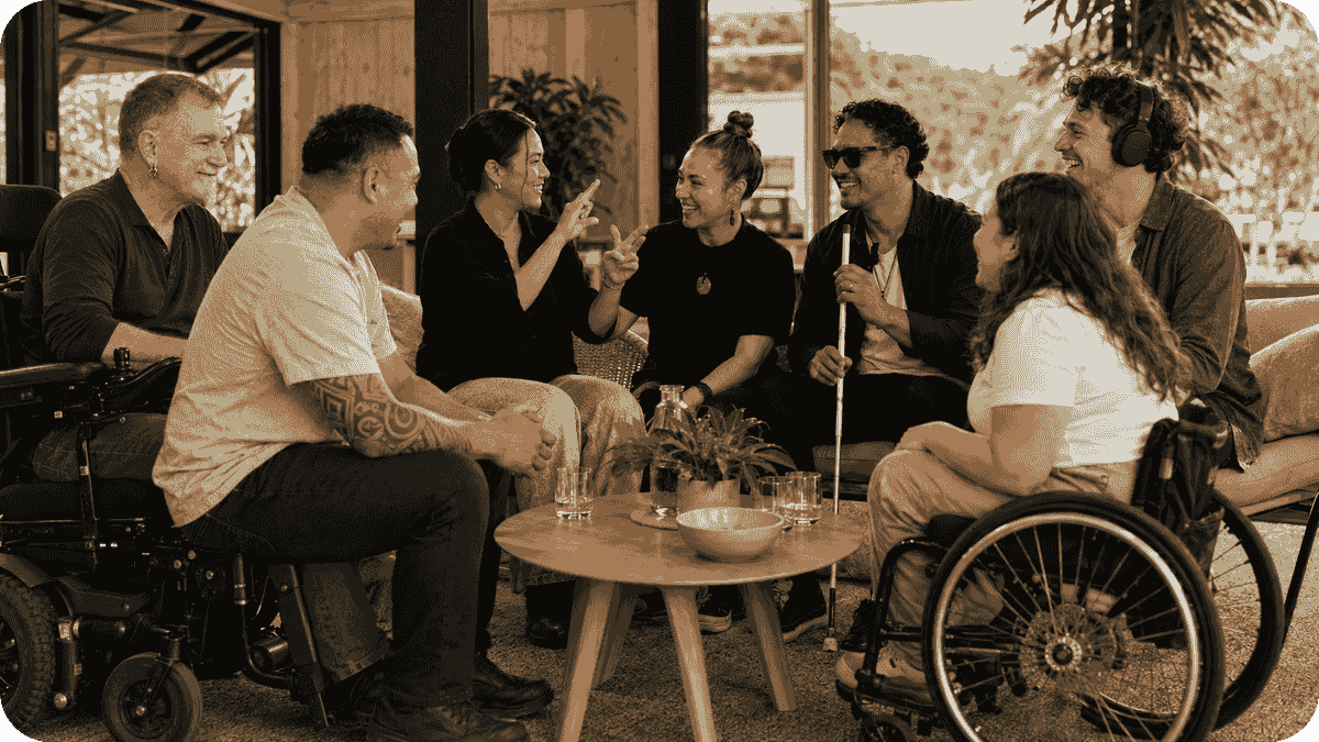 Pip, seated in a power wheelchair, joins six diverse people in a lively conversation around a table. The group includes a person signing, a man holding a white cane, a headphone wearer and a manual wheelchair user. Pip, seated in a power wheelchair, joins six diverse people in a lively conversation around a table. The group includes a person signing, a man holding a white cane, a headphone wearer and a manual wheelchair user.
