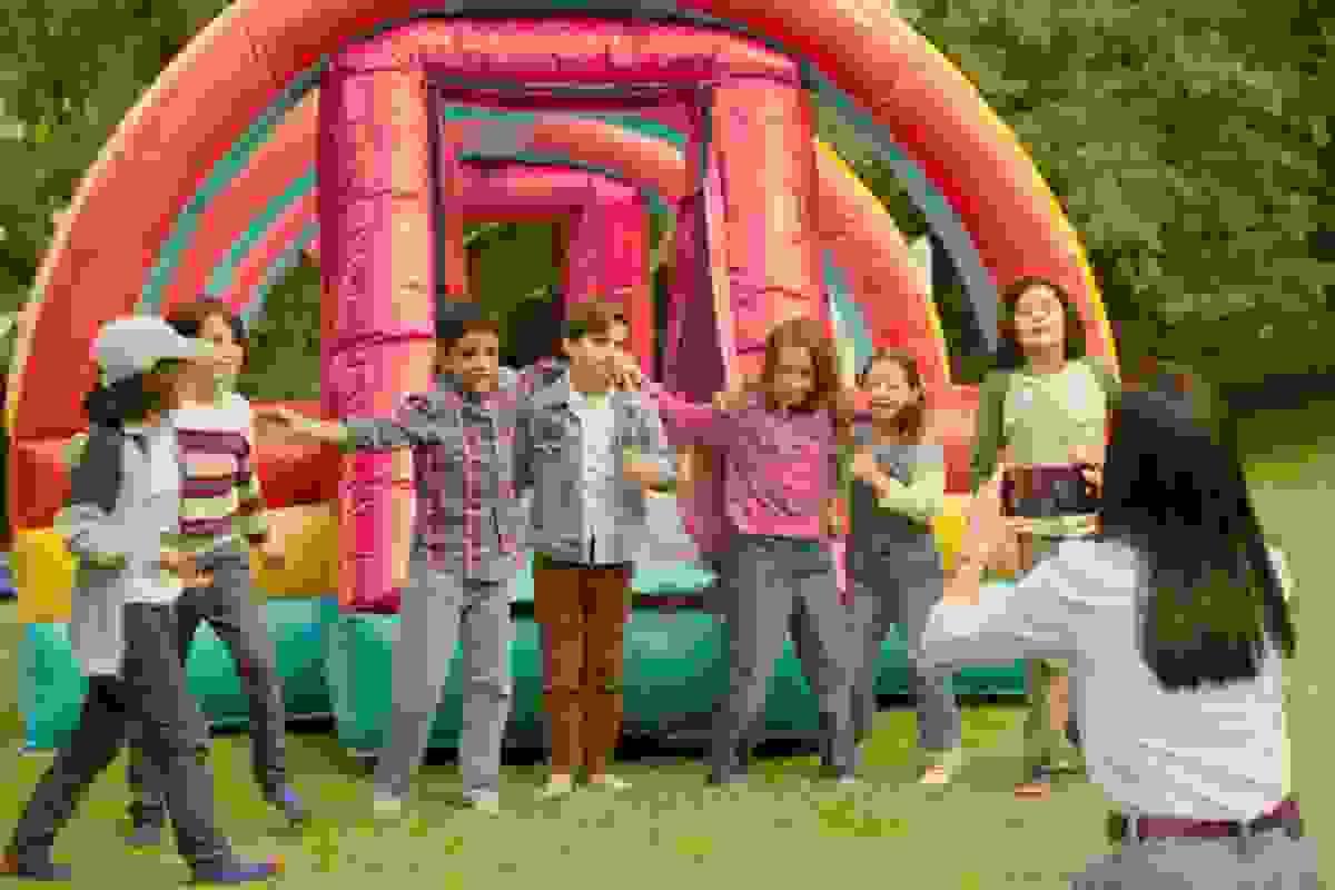 This image shows a group of children playing together in front of a colourful inflatable structure, with one of the adults in the group taking a photo of them. It captures a joyful and lively moment, with the children smiling and interacting in a playful manner.      