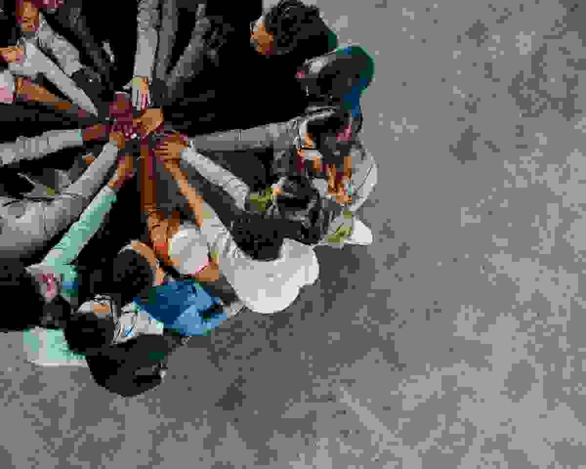 A diverse group of people stand in a circle with their hands stacked together in the center, viewed from above. The image emphasizes unity, teamwork, and collective effort against a grey carpeted background