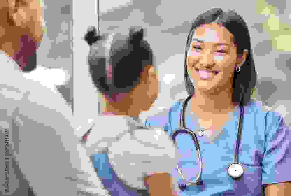 A healthcare professional in blue scrubs with a stethoscope around the neck speaks with a young girl who has her hair in two buns. The girl is accompanied by an adult, and they are in a bright room with large windows in the background.