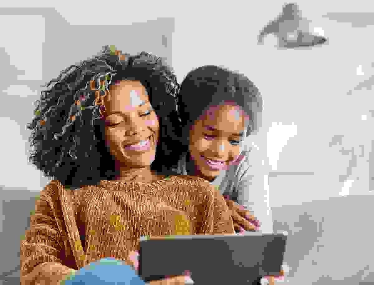 A mother sits on a couch wearing a mustard-colored sweater and holds a tablet, while her daughter leans over from behind in a light-colored shirt, both focused on the screen. A modern light fixture and greenery are visible in the background
