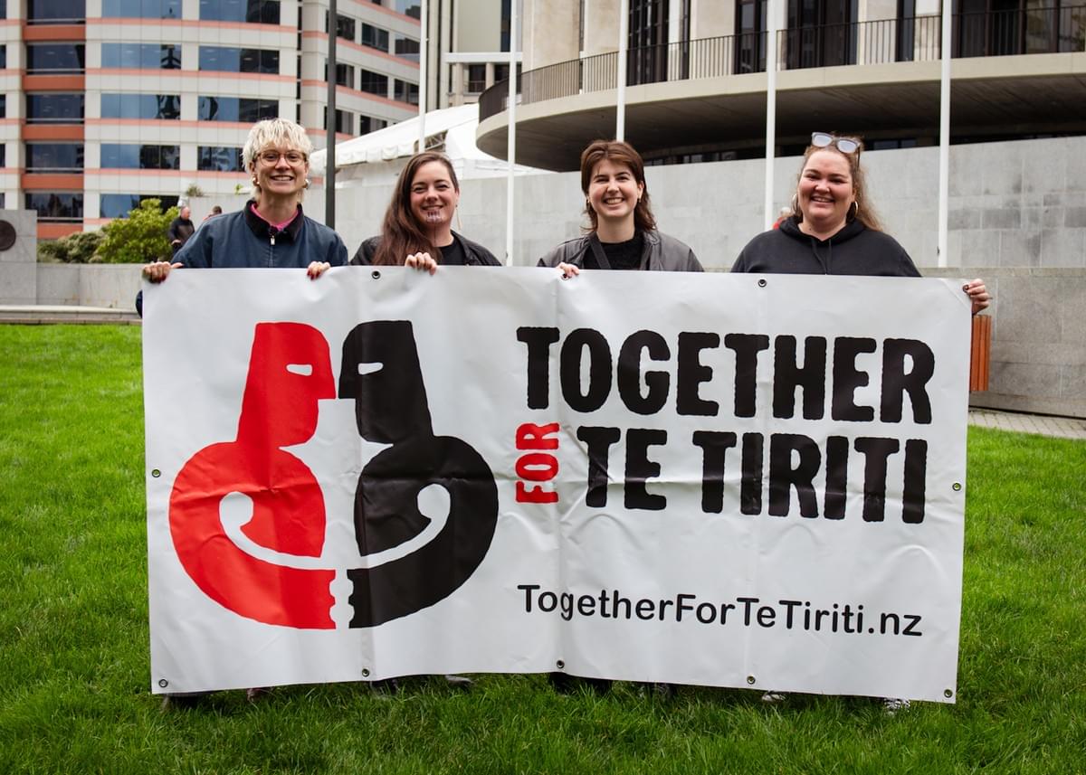 Hana, Kassie, Anastasia and Rangimarie stand smiling holding a giant Together for Te Tiriti banner outside Parliament.