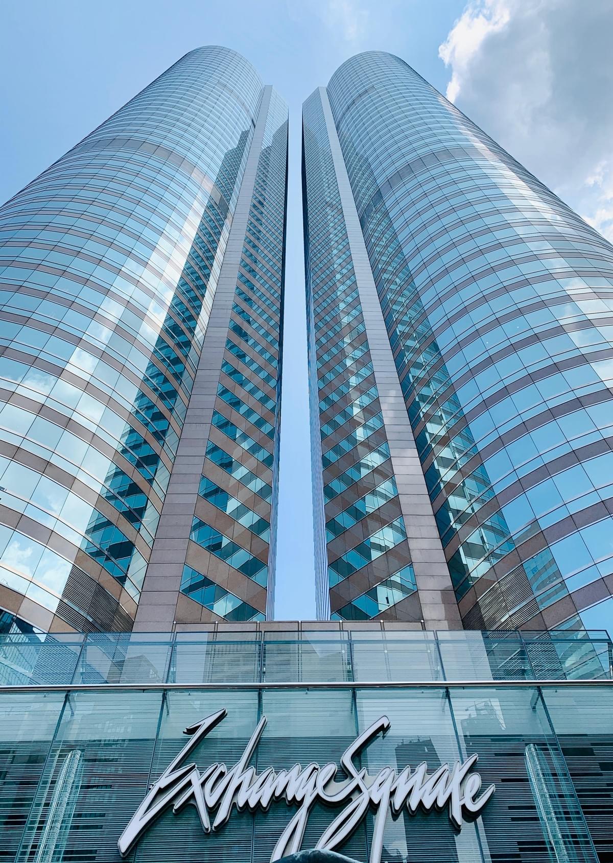 Looking up at the twin glass-clad towers of Exchange Square in Central, Hong Kong, with the cursive ‘Exchange Square’ entrance signage visible at street level