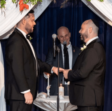 Officiant Schwa Potter performing ring exchange for two grooms under chuppah with unity candle Officiant Schwa Potter performing ring exchange for two grooms under chuppah with unity candle