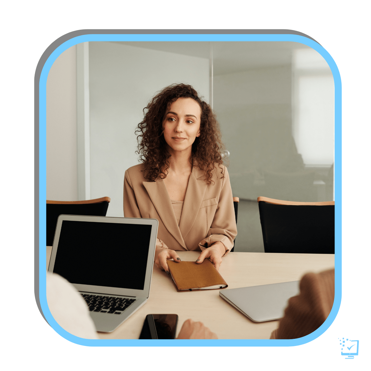 A young woman is sitting at a work desk across from two other professionals. She is framed in between them and their laptops.