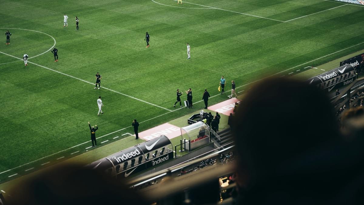 A high-angle view from the stadium stands showing a professional football match during a substitution; a coach is shouting tactical instructions to his players on the pitch while fans watch from the foreground.