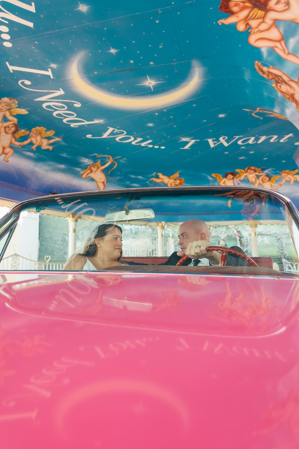 Couple in wedding attire posing inside the iconic vintage pink Cadillac at the Little White Wedding Chapel, featuring the unique floral overhead wallpaper, by Falling Rocks Photography.