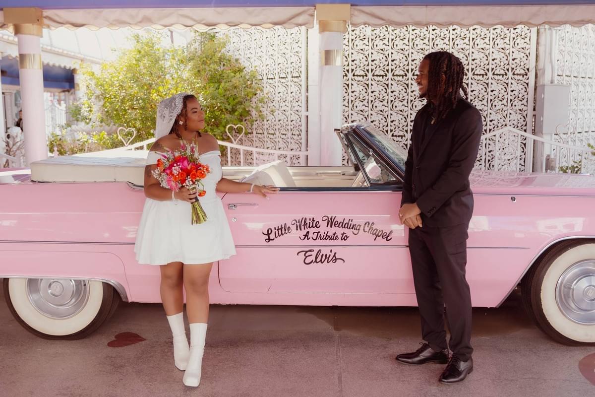 Newlyweds in wedding attire posing with the iconic vintage pink Cadillac at the Little White Wedding Chapel in Las Vegas.