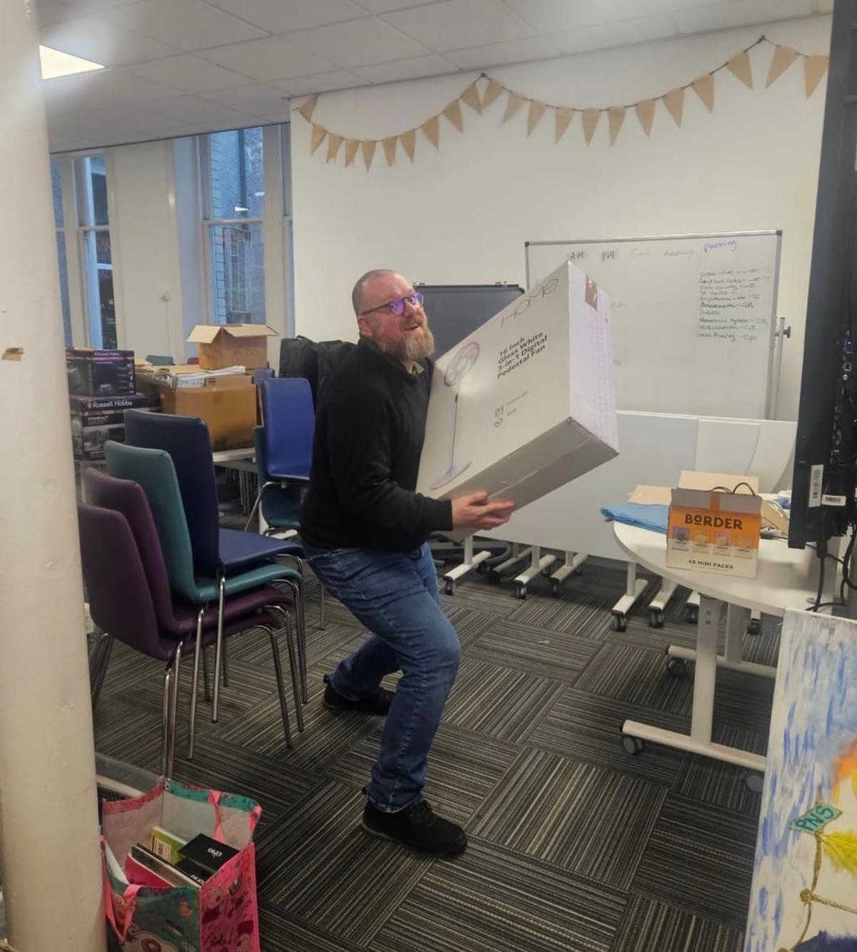 A person lifts a large boxed fan in an office meeting room with stacked chairs, boxes, and a whiteboard in the background