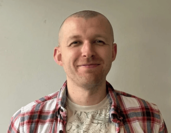 A person with short hair and a trimmed beard smiling warmly, wearing a light shirt and two necklaces, photographed against a plain light background. A person with short hair and a trimmed beard smiling warmly, wearing a light shirt and two necklaces, photographed against a plain light background.