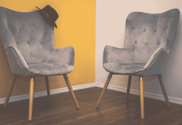 Two modern upholstered chairs with light grey fabric and wooden legs sit on a polished wooden floor in the corner of a room. The wall behind them is divided into two colours — a warm yellow on the left and soft white on the right — creating a calm, balanced contrast. A black wide‑brimmed hat rests casually on the back of the left chair, adding a touch of personality and warmth to the minimalist setting