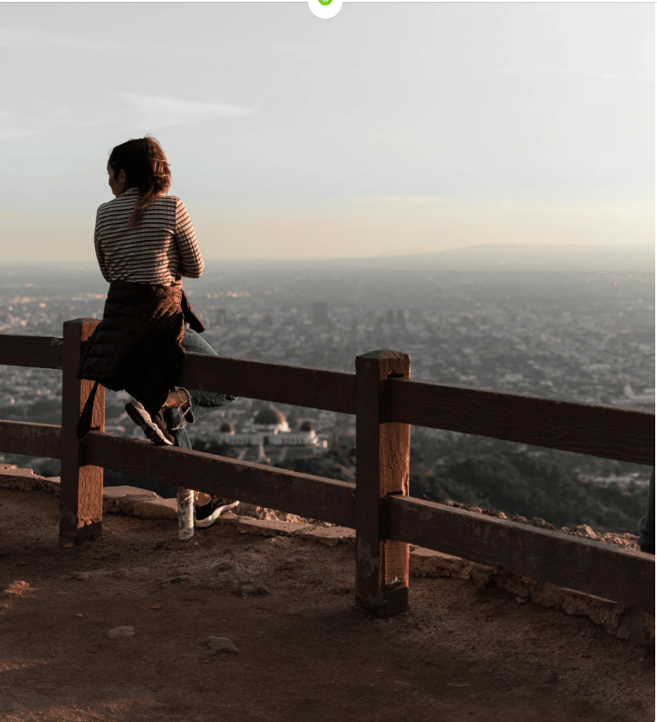 A casually dressed person sits alone on a wooden fence at a scenic overlook, gazing out over a sprawling cityscape below. The individual wears a striped shirt, jeans, and sneakers, with a dark jacket tied around their waist. A water bottle rests on the ground nearby, suggesting a pause during a walk or hike. The viewpoint is elevated—likely a hill or mountain—offering a panoramic view of densely packed urban buildings stretching toward the horizon. The sky above is hazy, casting a soft, muted light over the scene. The composition conveys a quiet moment of reflection and solitude, with the contrast between the natural foreground and the vast, structured city below evoking themes of contemplation, escape, and connection between nature and urban life